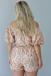 Woman wearing a tan and white romper that has off the shoulder short puffed sleeves, a cinched waistline, and a repeating leaf pattern indoors against a white wall. The photo shows the back of the romper. 