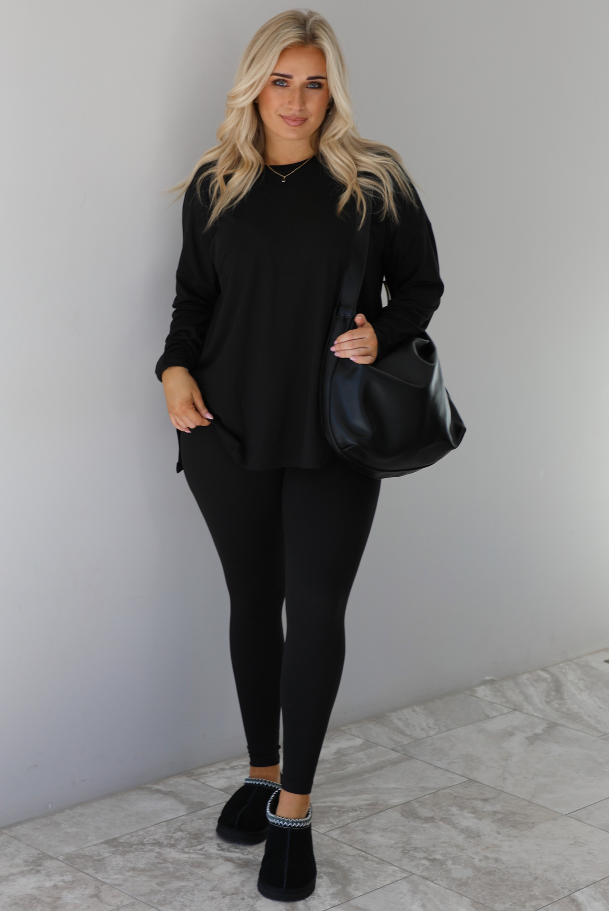Woman wearing a black two piece set that features an oversized long sleeved top and fitted leggings. The set is paired with a black vegan leather shoulder bag and black slide on slippers. The photo is taken indoors against a white wall with white marble flooring.