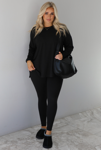 Woman wearing a black two piece set that features an oversized long sleeved top and fitted leggings. The set is paired with a black vegan leather shoulder bag and black slide on slippers. The photo is taken indoors against a white wall with white marble flooring.
