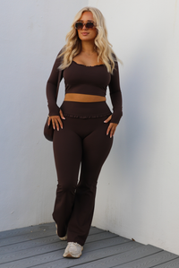 Woman model wearing a brown two-piece athleisure set. The top is cropped and the pants are high waisted. The photo is taken outdoors against a white wall.