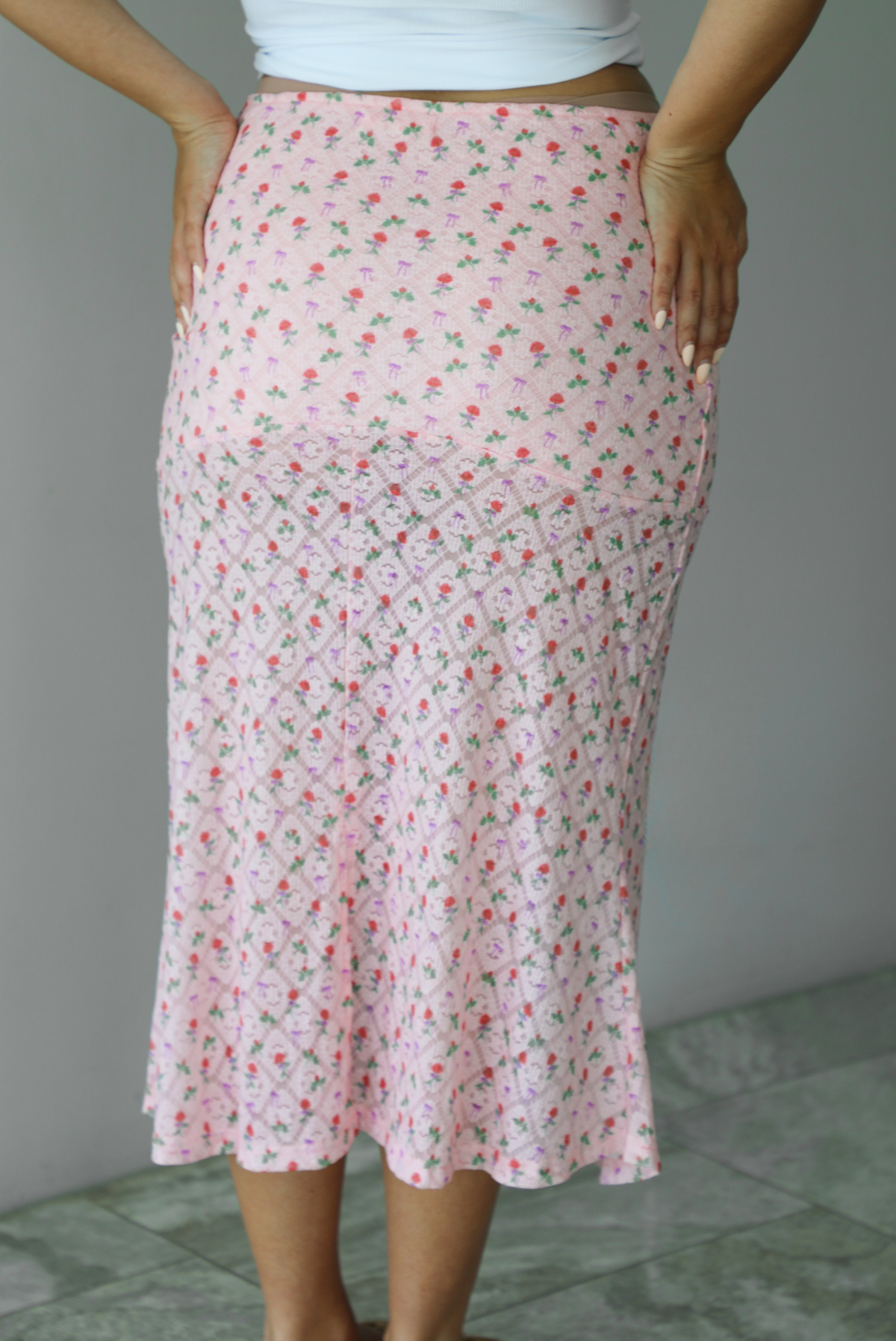 Woman wearing a pink midi length skirt with a multi-colored floral and bow repeating pattern indoors against a white wall with white marble flooring. The photo shows the back of the skirt. 