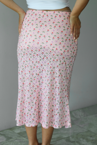 Woman wearing a pink midi length skirt with a multi-colored floral and bow repeating pattern indoors against a white wall with white marble flooring. The photo shows the back of the skirt. 