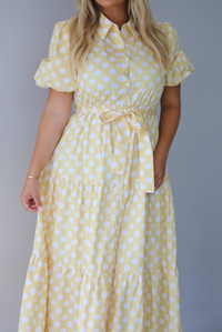 A woman wearing a yellow and white polka dot midi dress with puff sleeves and a tie waist and functional button closures along the front, standing indoors against a white wall.