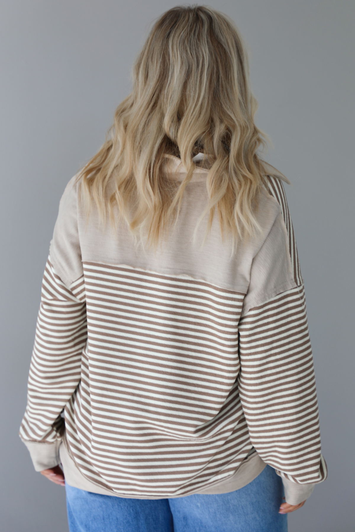 Woman wearing a mocha and cream striped pullover with a collared neckline, quarter button closures in the front, that has an oversized fit. The photo shows the back of the sweater. The photo is taken indoors against a white wall.