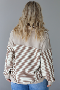 Woman wearing a mocha and cream striped pullover with a collared neckline, quarter button closures in the front, that has an oversized fit. The photo shows the back of the sweater. The photo is taken indoors against a white wall.
