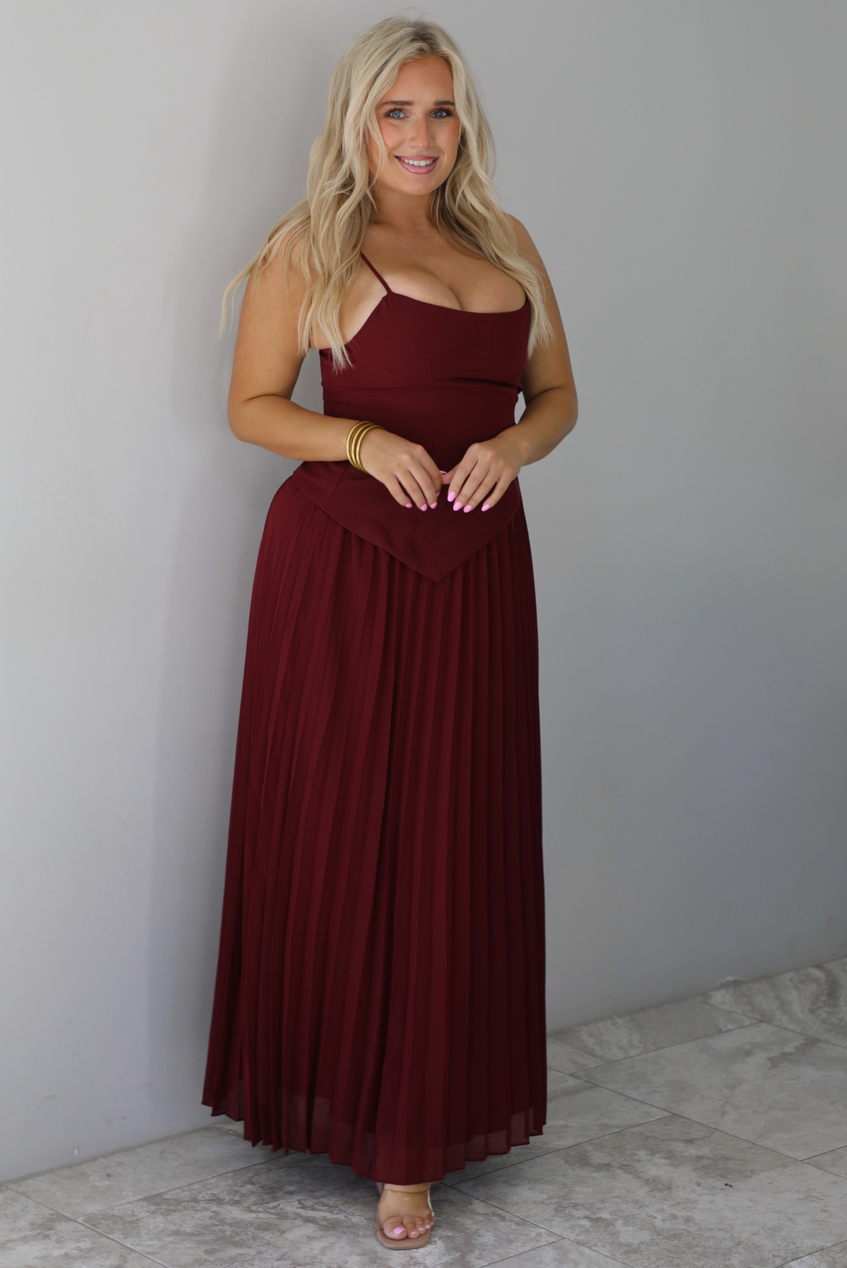 A woman wearing a wine colored formal maxi dress with a fitted, drop waist design with a crimped floral maxi skirt, standing indoors against a white wall with white marble flooring. 
