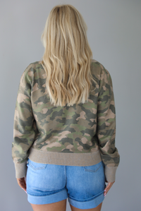 Woman wearing a camo cardigan with functional button closures in the front tucked into high waisted denim shorts. The photo is taken in front of a white wall and  shows the back of the cardigan. 