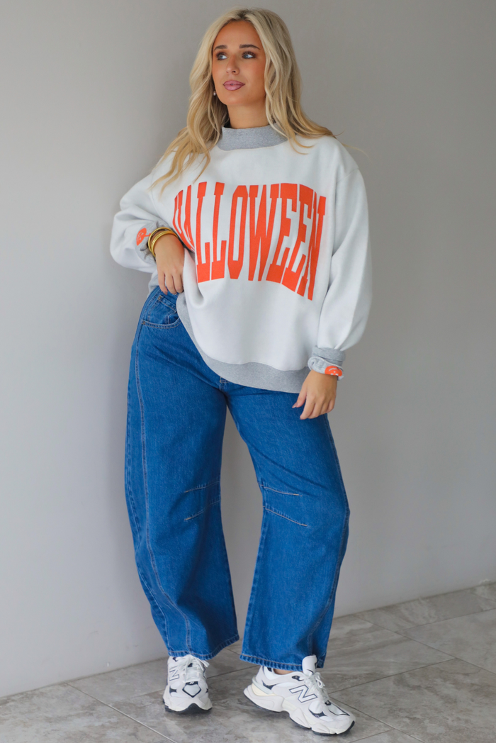 A woman wearing a light grey and dark grey trim oversized sweatshirt that says "HALLOWEEN" in collegiate font on the front, with jack-o-lantern details on the rolled up sleeves, paired with dark wash denim jeans, standing indoors against a white wall with white marble flooring. 