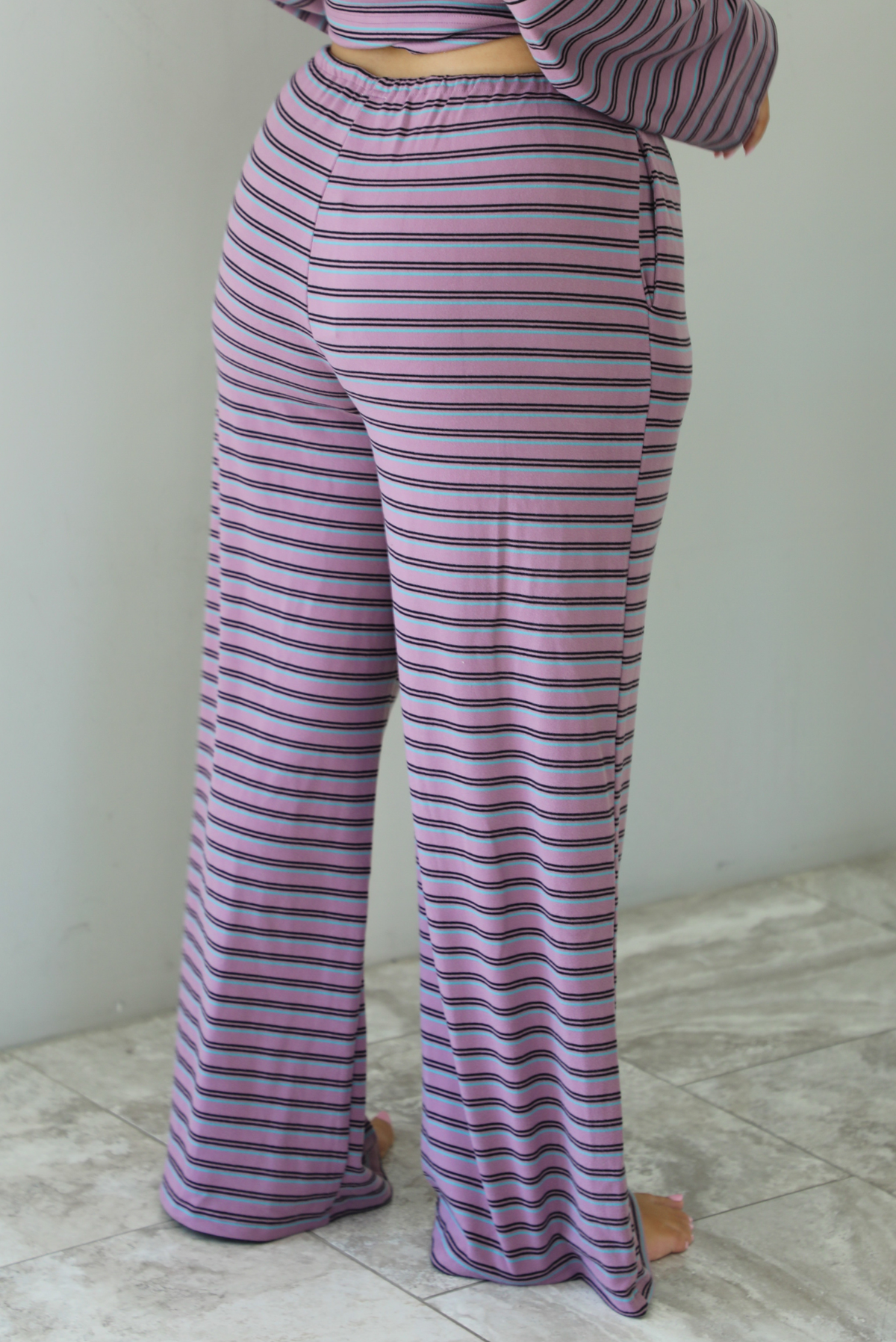 A woman shown from the waist down from behind wearing a purple matching set with a black and blue striped pattern featuring an oversized long sleeved top and high waisted flared hem pants, standing indoors against a white wall with white marble flooring. The photo shows a closeup of the back of the pants. 