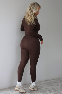 The photo features a model wearing a brown fitted jumpsuit with long sleeves that have thumb holes, a rounded neckline with functional button closures, a ribbed texture, and is super stretchy. The jumpsuit is paired with white scrunchie socks and beige sneakers. The photo is taken indoors against a white wall with white marbled flooring, and shows the back of the jumpsuit. 