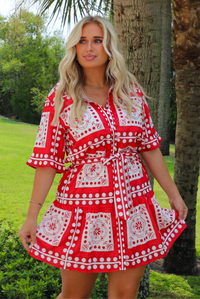 A woman wearing a red and white patterned short dress stands outdoors in front of a tree, with grass and greenery in the background.