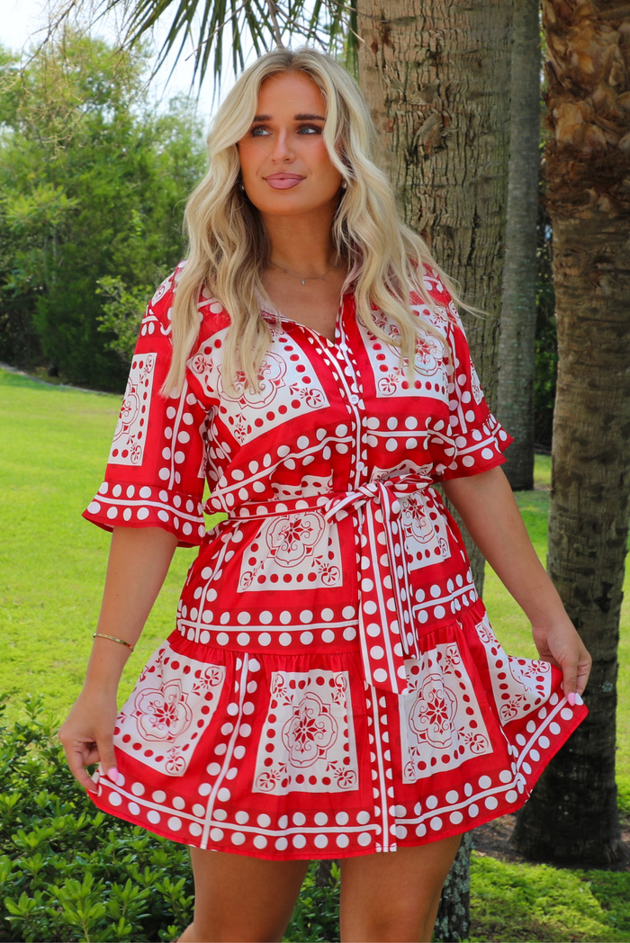 A woman wearing a red and white patterned short dress stands outdoors in front of a tree, with grass and greenery in the background.
