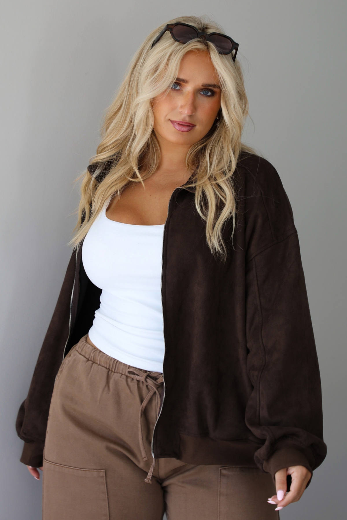 A woman wearing a dark brown suede jacket with a white tank and mocha colored mid-to-low rise pants, standing indoors against a white wall.