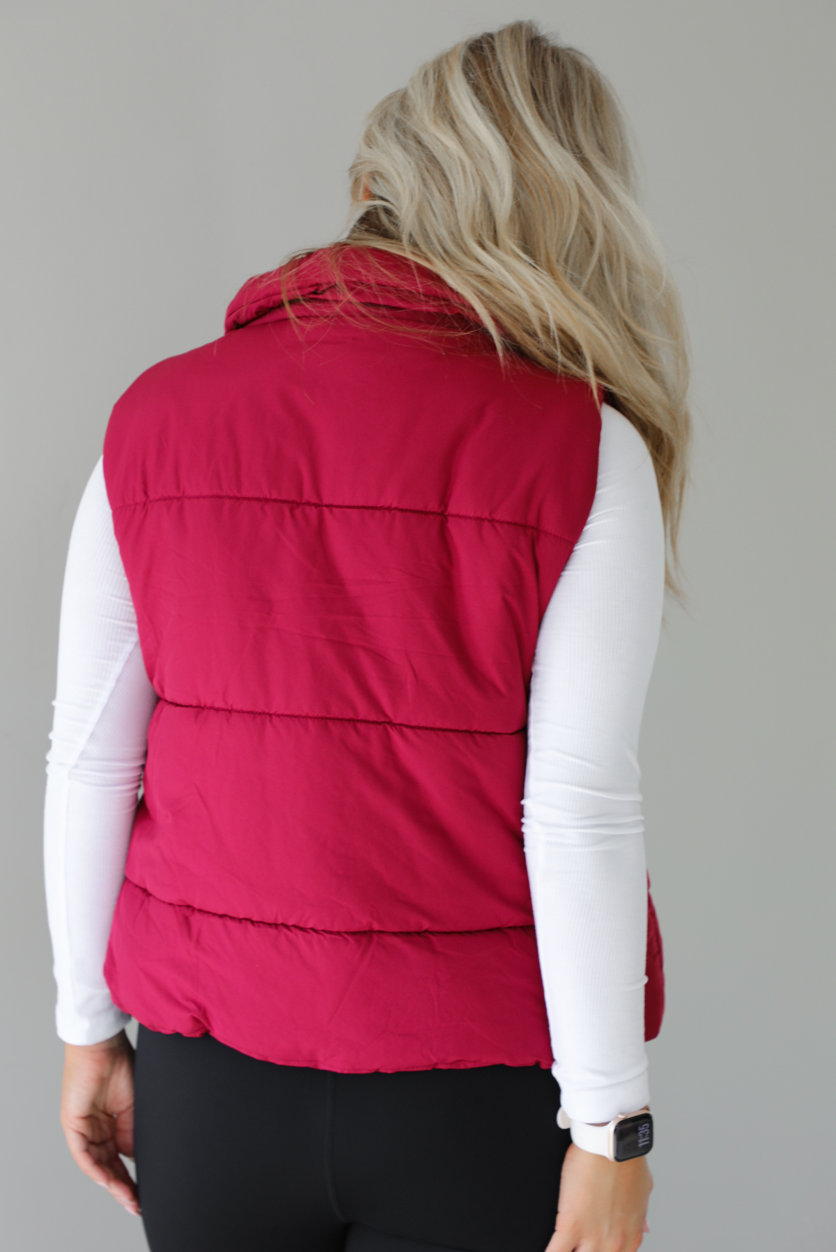 Woman wearing a wine red puffer vest, white long-sleeve shirt, and black leggings indoors against a white wall. The puffer vest has a collared design, functional button closures at the neckline, an unzipped closure in the front, and functional pockets on either side. The photo shows the back of the puffer vest.