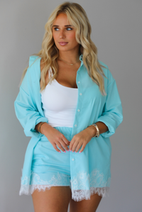 A woman stands indoors against a white wall wearing a light aqua blue matching lounge set consisting of a long open-front button-down and shorts with white lace trim, over a white tank top.