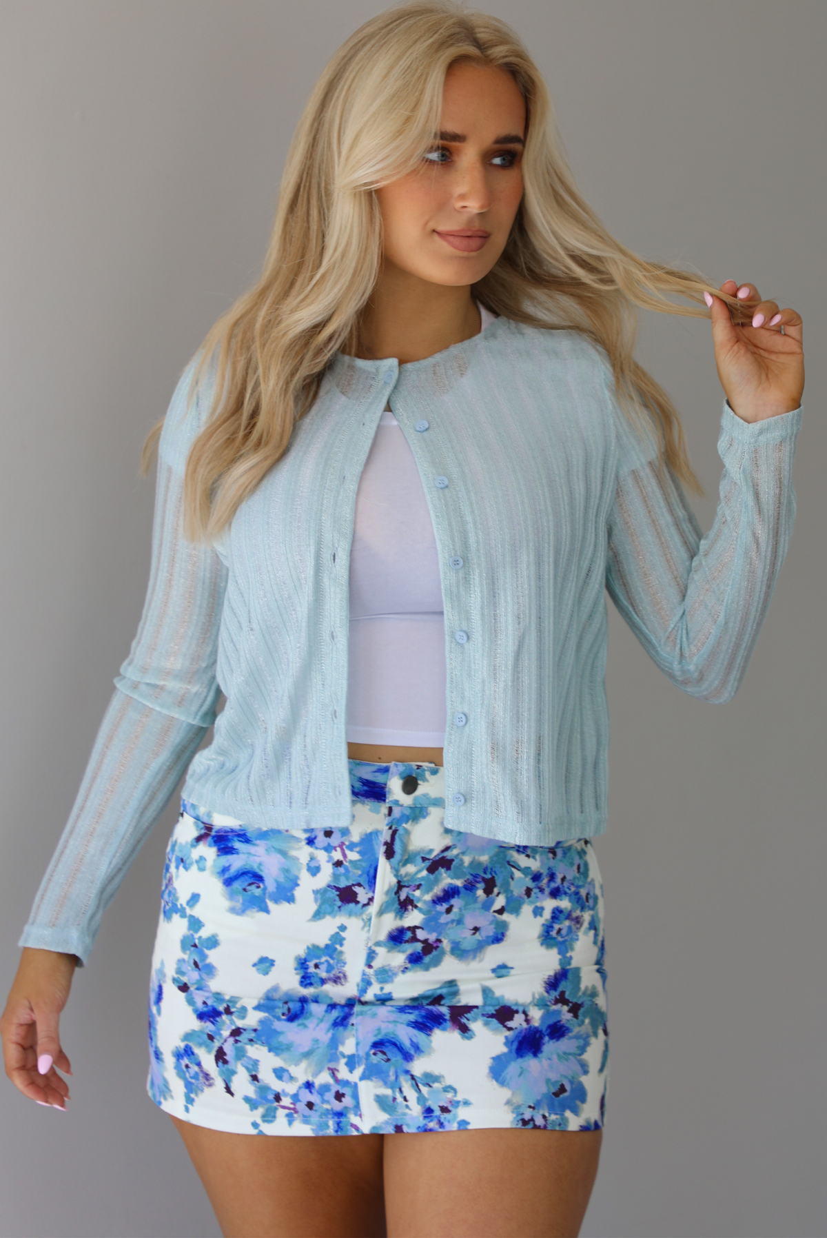Woman wearing a light blue cardigan that is buttoned at the top button only with a white undershirt and a white and blue floral denim mini skirt indoors against a white wall.