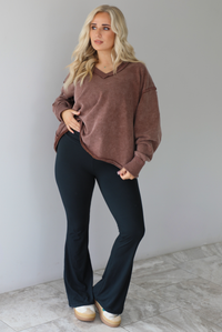 Woman wearing a mauve oversized v-neck sweater with raw hem details with high waisted black flared leggings. The photo is taken in front of a white wall with white marble flooring. 