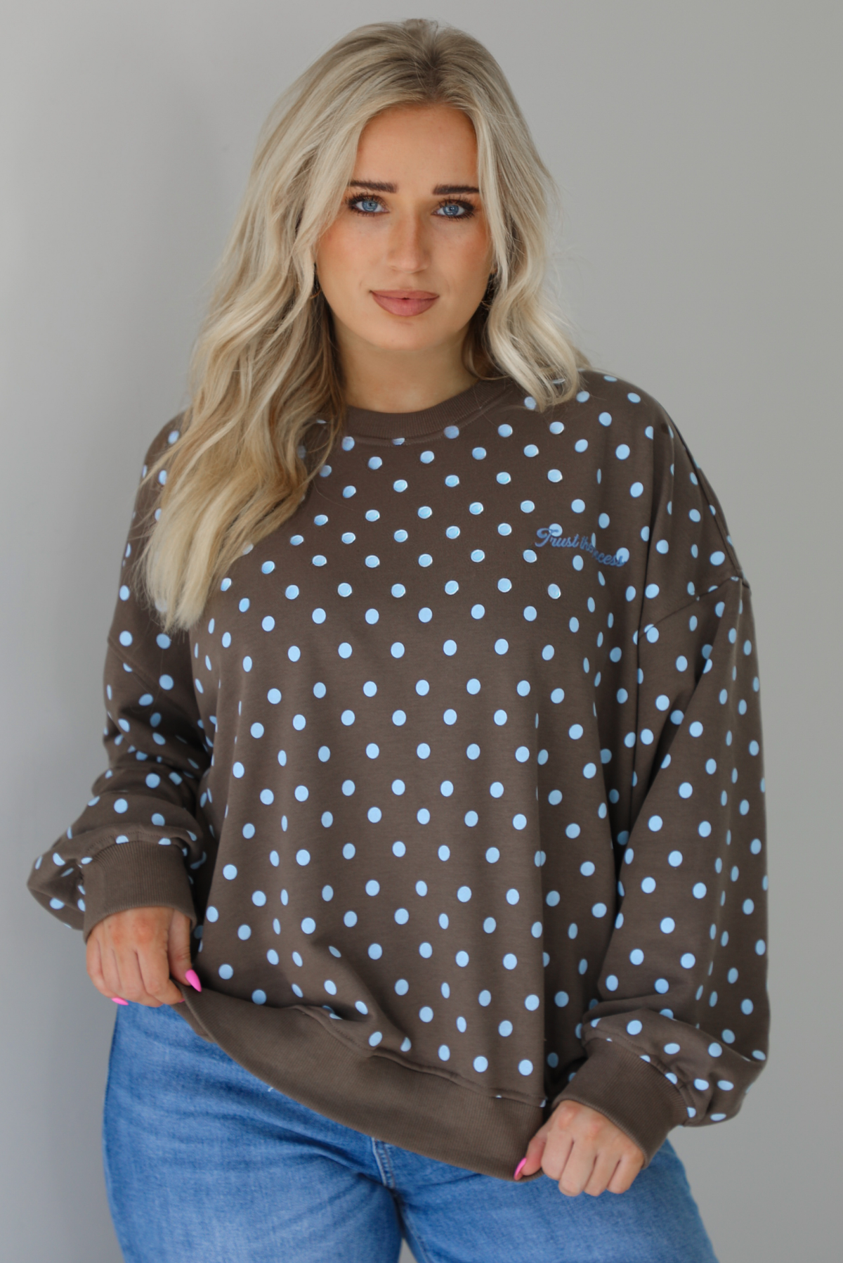 Woman wearing a brown sweater that has a blue polka dot print pattern throughout with blue embroidered writing on the left side that reads 'Trust The Process.' The sweater is paired with a medium wash denim. The photo shows a closeup of the sweater, which is untucked to show the full length. The photo is taken indoors against a white wall.