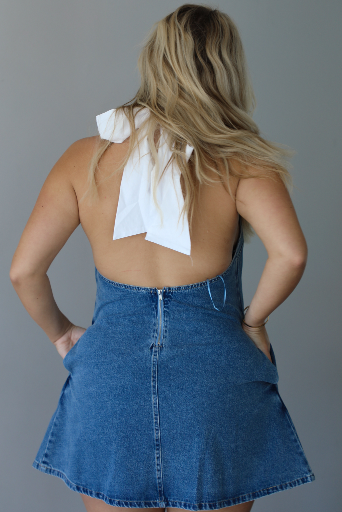 Woman wearing a denim blue dress indoors against a white wall. The photo shows a closeup of the back of the dress, which has a halter tie neckline, an open back, and a zipper closure. 