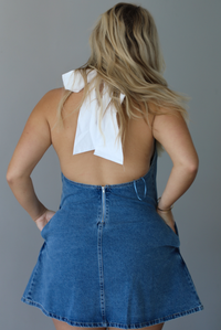 Woman wearing a denim blue dress indoors against a white wall. The photo shows a closeup of the back of the dress, which has a halter tie neckline, an open back, and a zipper closure. 