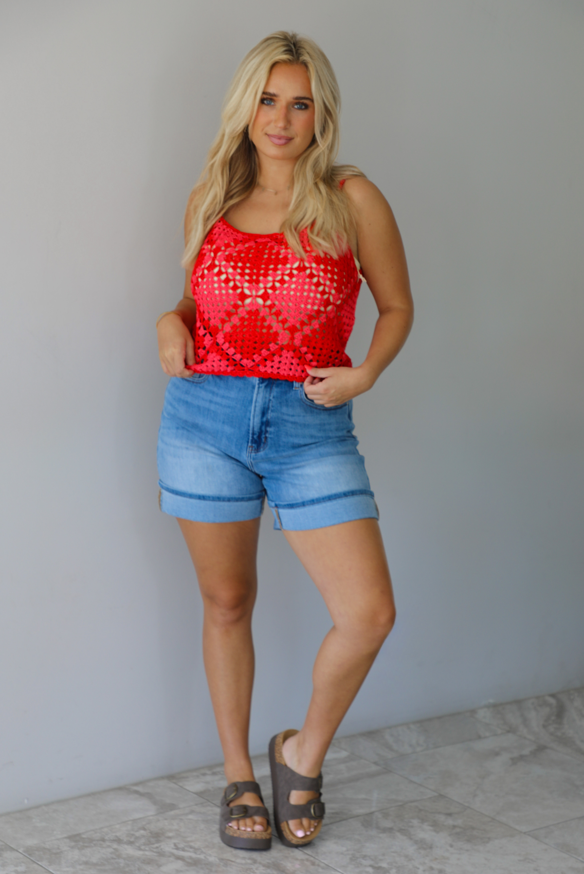 Woman wearing a pink and red loose knit crochet cropped top with light wash denim shorts indoors against a white wall with white marble flooring. 