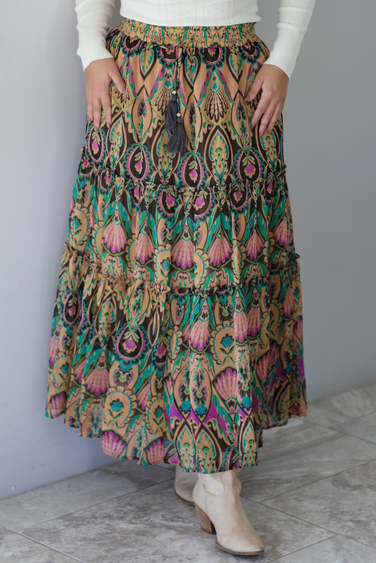 A woman, whose face is not showing, wearing a long sleeved cream top and multi-colored maxi length skirt paired with knee high sand colored ankle boots, standing indoors against a white wall with white marble flooring. 