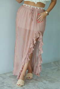 Woman wearing a cream cropped, off the shoulder top with a high waisted blush maxi skirt indoors against a white wall with white marble flooring. The photo shows a close-up of the skirt, showing how it has an elastic and drawstring waistline and a split hem design that has a ruffled detail.