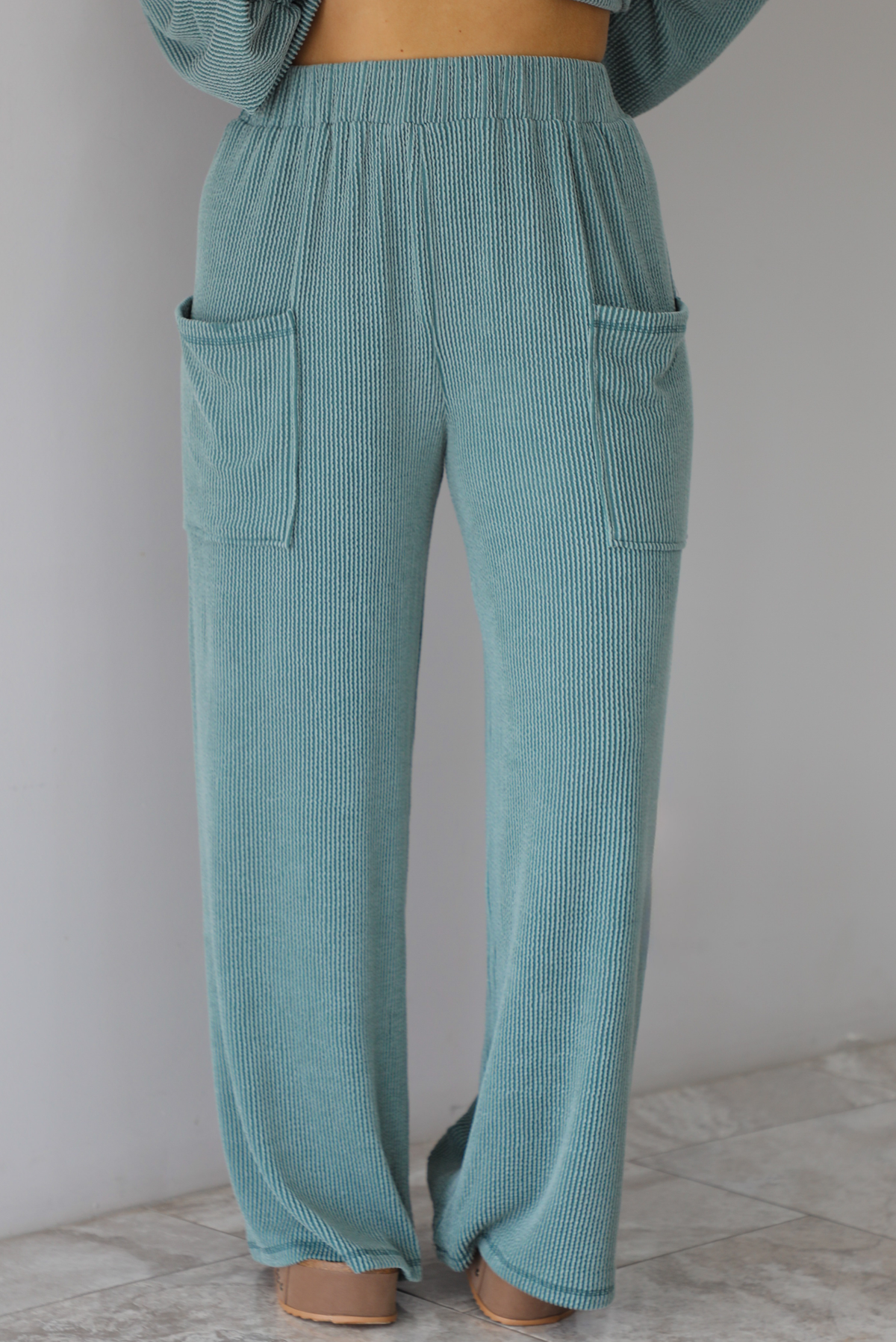 Woman wearing a teal two piece set that has a v-neck long sleeved oversized top and high waisted pants. The photo shows a closeup of the pants of the set, which have an elastic waistline, functional pockets on either side, with a ribbed texture. The photo is taken indoors against a white wall with white marble flooring. 