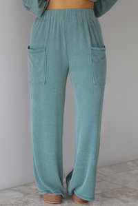 Woman wearing a teal two piece set that has a v-neck long sleeved oversized top and high waisted pants. The photo shows a closeup of the pants of the set, which have an elastic waistline, functional pockets on either side, with a ribbed texture. The photo is taken indoors against a white wall with white marble flooring. 