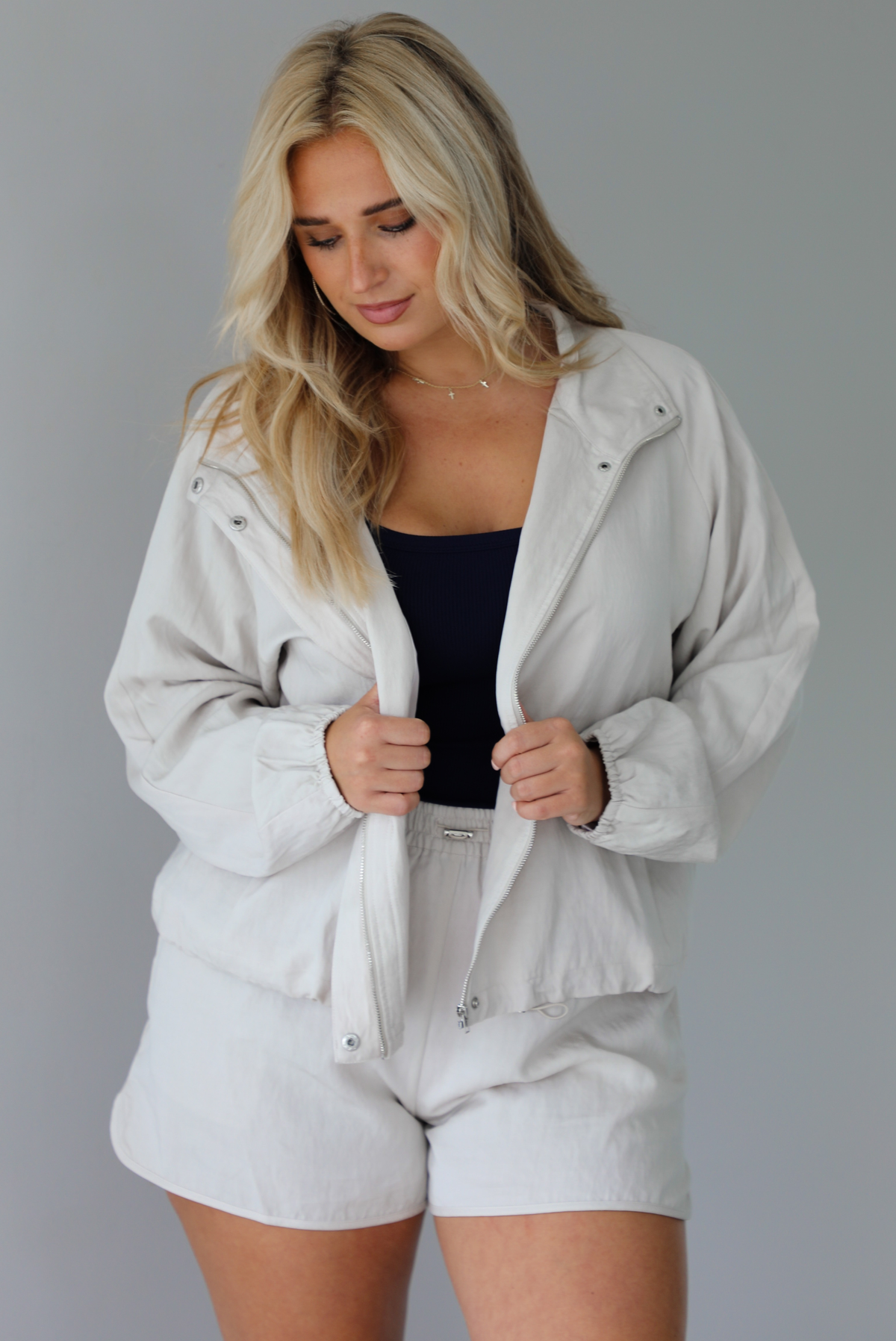 Woman wearing a stone colored two piece set featuring a zip up jacket and shorts with a navy tank tucked into the shorts indoors against a white wall.