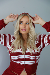 A woman modeling a matching red and white striped knit set with the hoodie on the top up, has a front zipper, drawstring hood, standing indoors against a white wall.