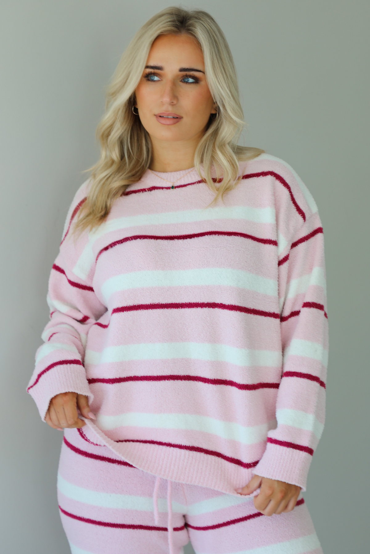 Woman wearing a pink set with a darker pink and white striped design. The set consists of an oversized sweater top and high waisted pants. The photo shows a closeup of the sweater, untucked. The photo is taken indoors against a white wall. 