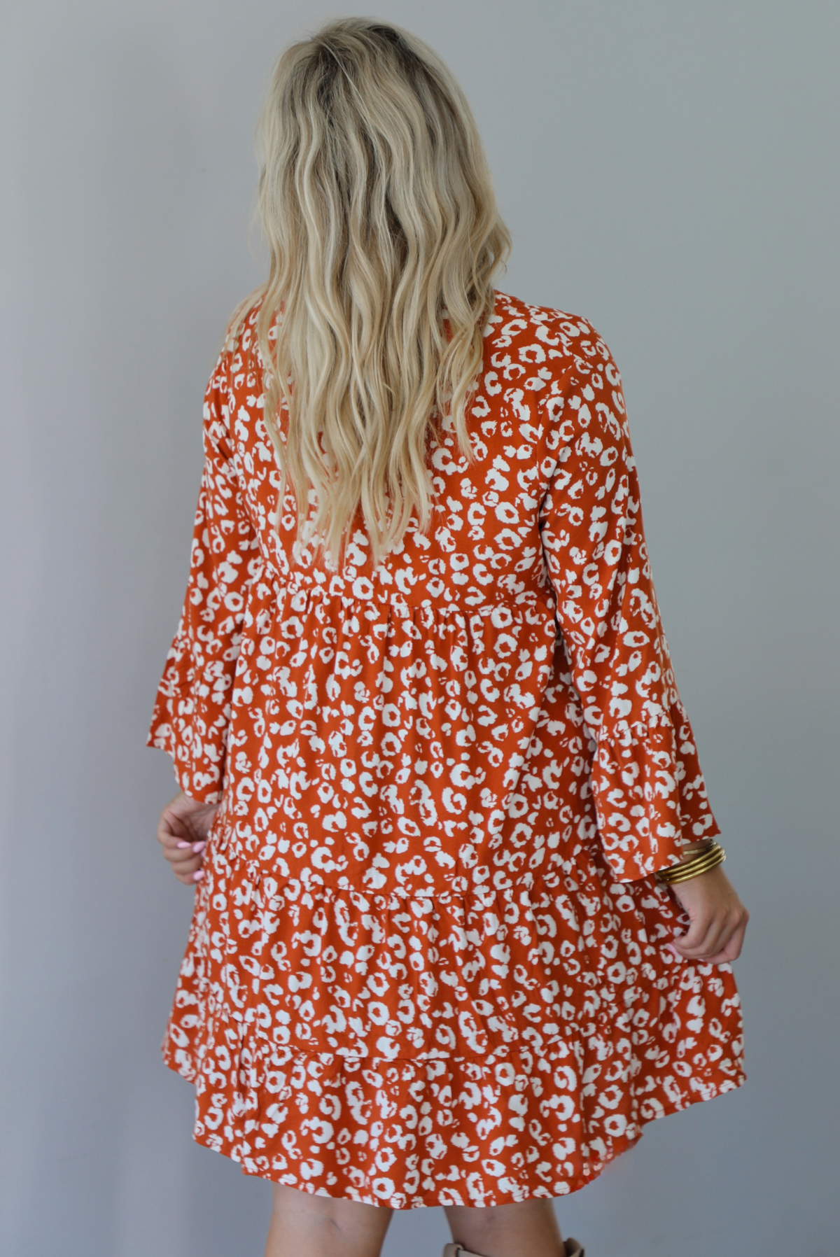 A woman shown from behind wearing an orange and white animal print long sleeved, flowy mini dress, standing indoors against a white wall.