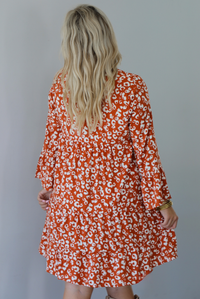 A woman shown from behind wearing an orange and white animal print long sleeved, flowy mini dress, standing indoors against a white wall.