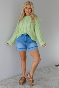 Woman wearing a green, long sleeved sweater with a loose knit design, functional button closures in the front, cuffed sleeves, and a raw, unfinished hem with medium wash denim shorts indoors against a white wall with white marble flooring. 