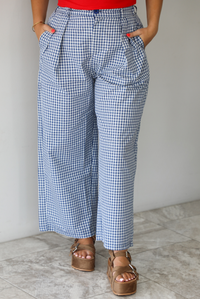 Woman wearing a navy and white plaid print pants that have a high waist, pleated details, functional pockets on either side, and a professional fit indoors against a white wall with white marble flooring. 