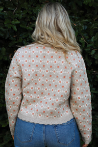 Woman model wearing a grey cardigan that has an orange and white floral print pattern throughout, long sleeves, an asymmetrical hem, and functional button closures in the front, with a long sleeved white top, and a pair of medium wash denim jeans outdoors in front of a green hedge. The photo shows a close up of the back of the cardigan. 