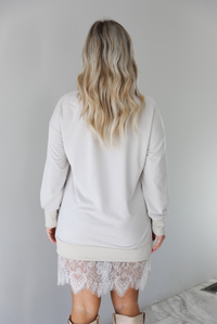 Woman wearing a sand colored sweater dress that has long sleeves, a relaxed fit, and a lace detailing at the hem. The photo is taken indoors against a white wall and shows a closeup of the back of the dress.