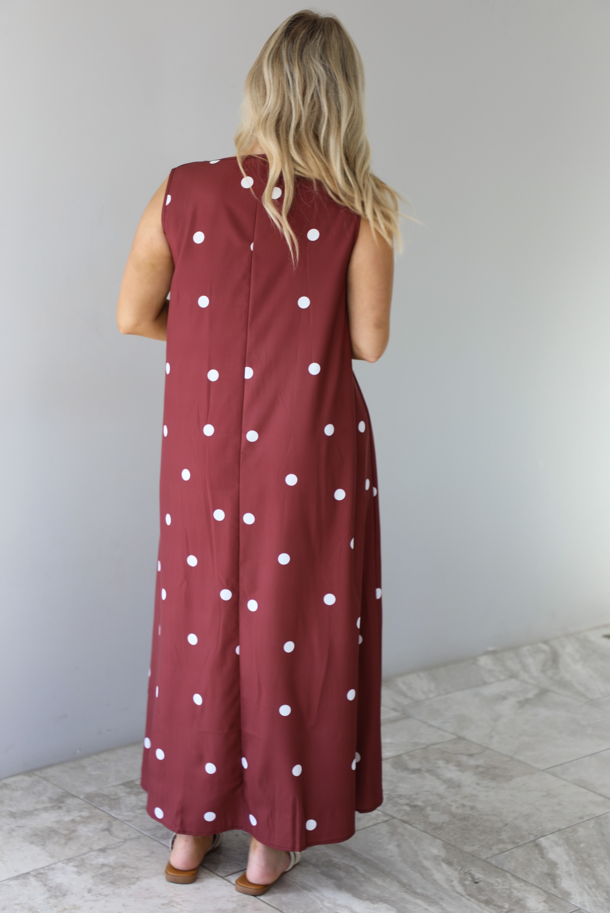 Woman wearing a red brown maxi length dress that has a white polka dot print pattern throughout. The photo shows the back of the dress. The photo is taken indoors against a white wall with white marble flooring. 