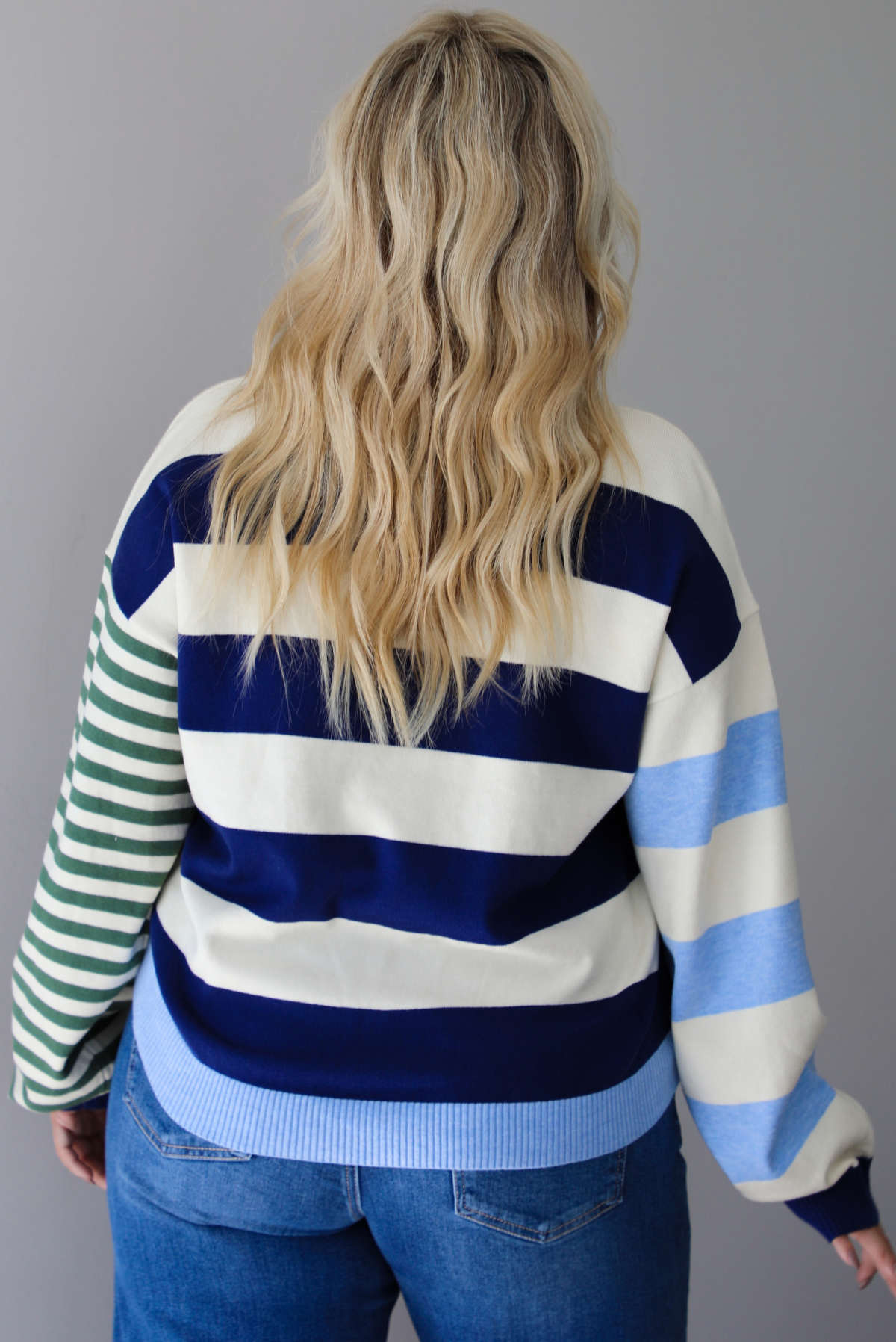Woman shown from behind wearing a navy, cream, blue, and green alternating striped design throughout, paired with dark denim jeans, standing indoors against a white wall.