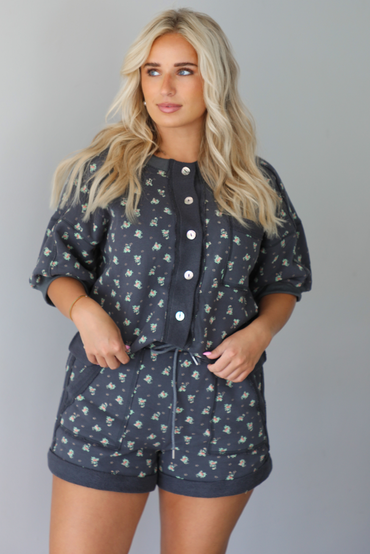Woman wearing a charcoal with a multi-colored floral two piece set featuring an oversized top with puffed sleeves and shorts indoors against a white wall. The photo shows a close up of the set, showing the functional button closures on the front of the top and the elastic and drawstring waistline on the shorts. 