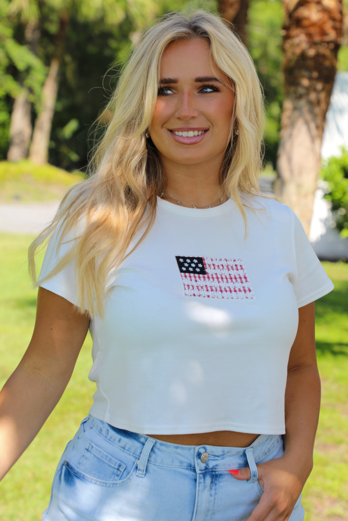 Woman wearing a white baby tee with an embroidered American Flag on the front with light wash denim shorts outdoors with greenery and palm trees in the background. 