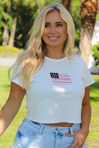 Woman wearing a white baby tee with an embroidered American Flag on the front with light wash denim shorts outdoors with greenery and palm trees in the background. 