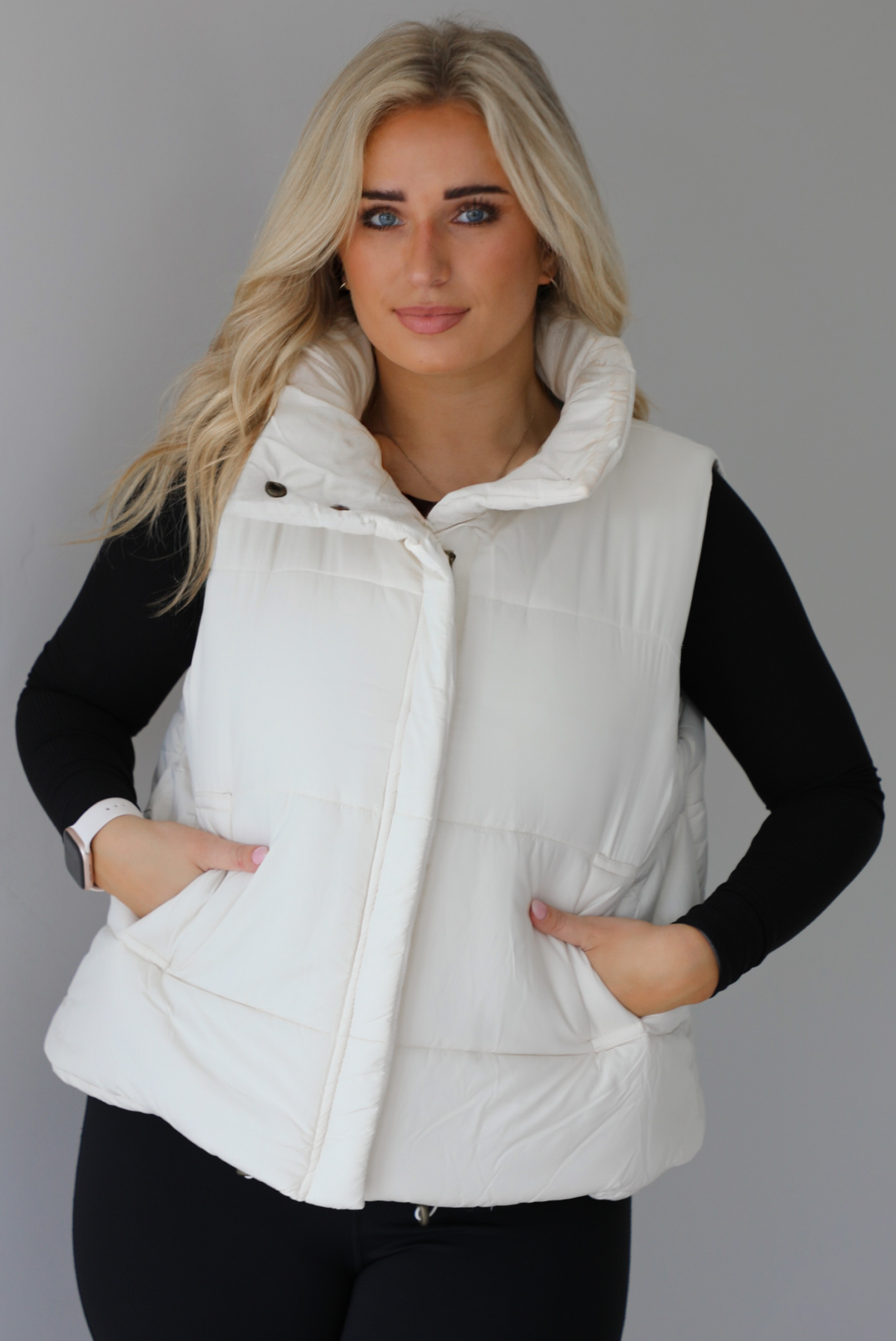 Woman wearing a light sand colored puffer vest with a fitted black long sleeved top and fitted leggings. The vest has functional button closures at the top, a zipper closure in the front, and functional pockets on either side. The photo shows the vest zipped up. The photo is taken indoors against a white wall with white marble flooring. 
