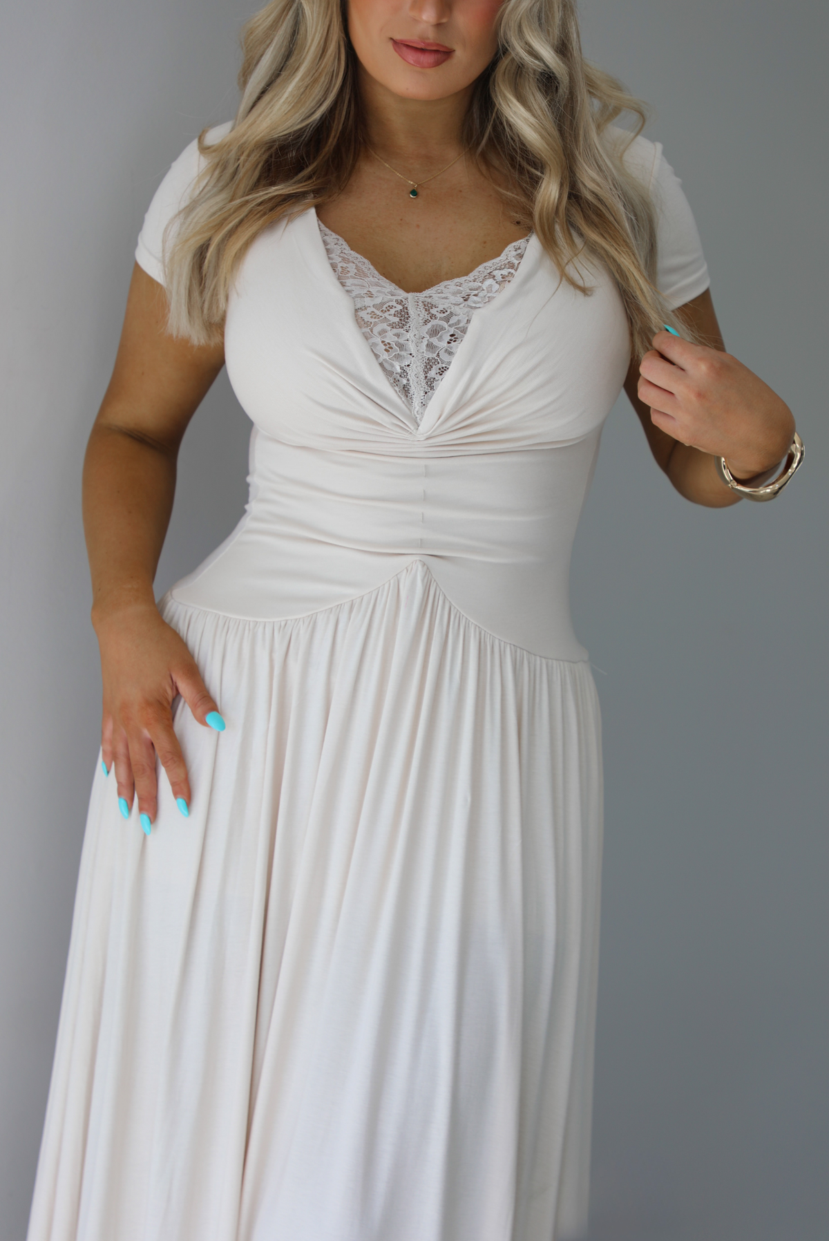 Woman wearing a light sand colored maxi length dress that has a fitted bodice with lace detailing in the center of the bust, a drop waist design, short sleeves, and a flowy skirt. The photo is taken indoors against a white wall and shows a closeup of the bodice of the dress. 
