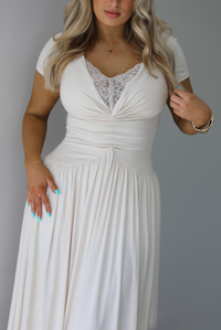 Woman wearing a light sand colored maxi length dress that has a fitted bodice with lace detailing in the center of the bust, a drop waist design, short sleeves, and a flowy skirt. The photo is taken indoors against a white wall and shows a closeup of the bodice of the dress. 