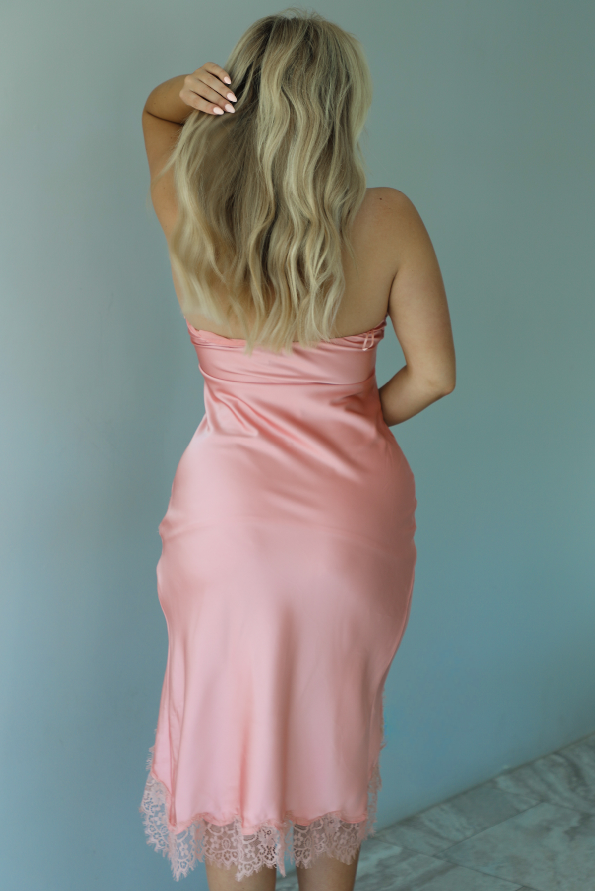 Woman wearing a strapless, peach satin midi dress with a split hen design and lace details on the hem indoors against a white wall and shows the back of the dress. 
