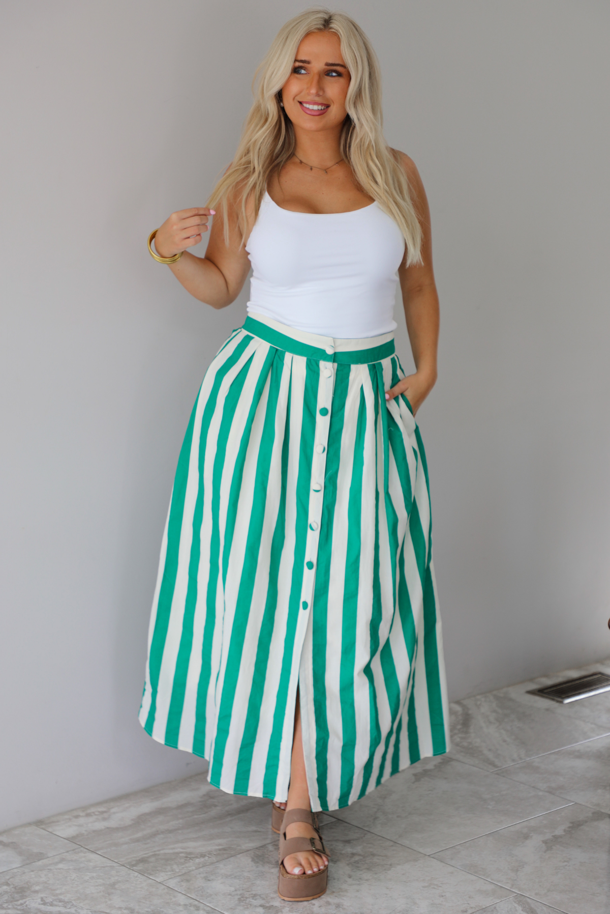 A woman wearing a white tank top and a maxi length cream and green striped skirt,  which has a high waistline, functional pockets on either side, functional button closures in the front, and has a flowy fit, standing indoors against a white wall with white marble flooring. 