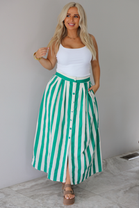 A woman wearing a white tank top and a maxi length cream and green striped skirt,  which has a high waistline, functional pockets on either side, functional button closures in the front, and has a flowy fit, standing indoors against a white wall with white marble flooring. 