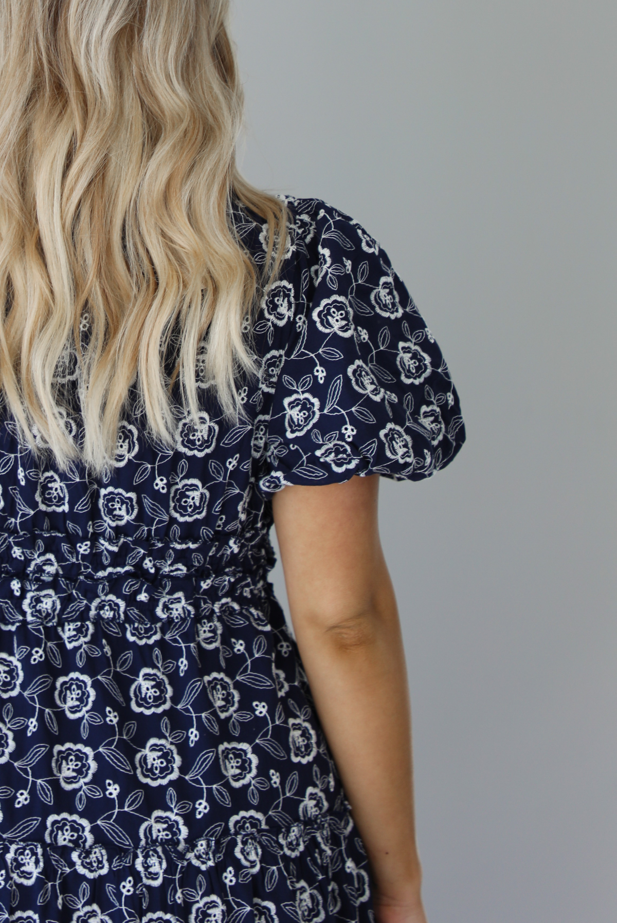 Woman wearing a navy dress that has a white embroidered floral print pattern throughout, a cinched waistline, and flowy maxi length skirt. The photo shows a closeup of the puffed sleeve and is taken in front of a white wall. 